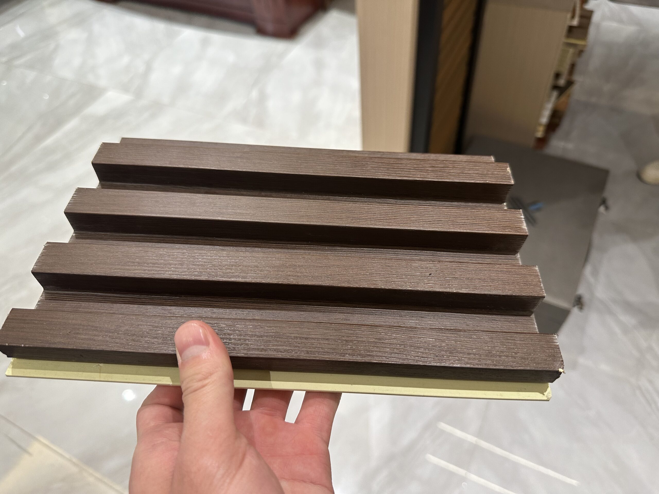 Dark brown grooved decorative wall panel sample with a textured wood-look finish held by hand.